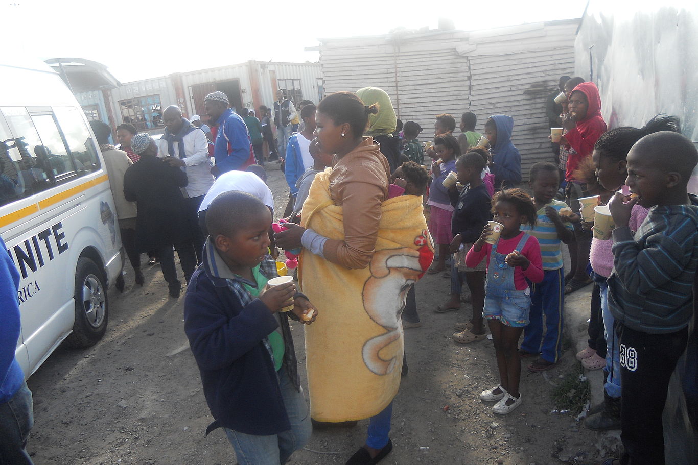 Kinder und Erwachsene stehen bei einem Bus von Africa Unite. Einige trinken aus Pappbechern.