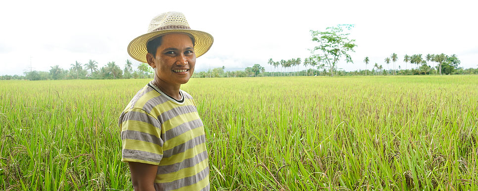 Ein Bauer mit Hut lächelt in die Kamera und steht auf einem riesigen Reisfeld.
