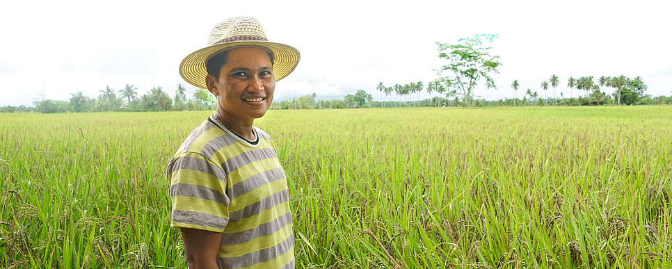 Ein Bauer auf der philippinischen Insel Mindanao Ein Bauer mit Hut lächelt in die Kamera und steht auf einem riesigen Reisfeld.