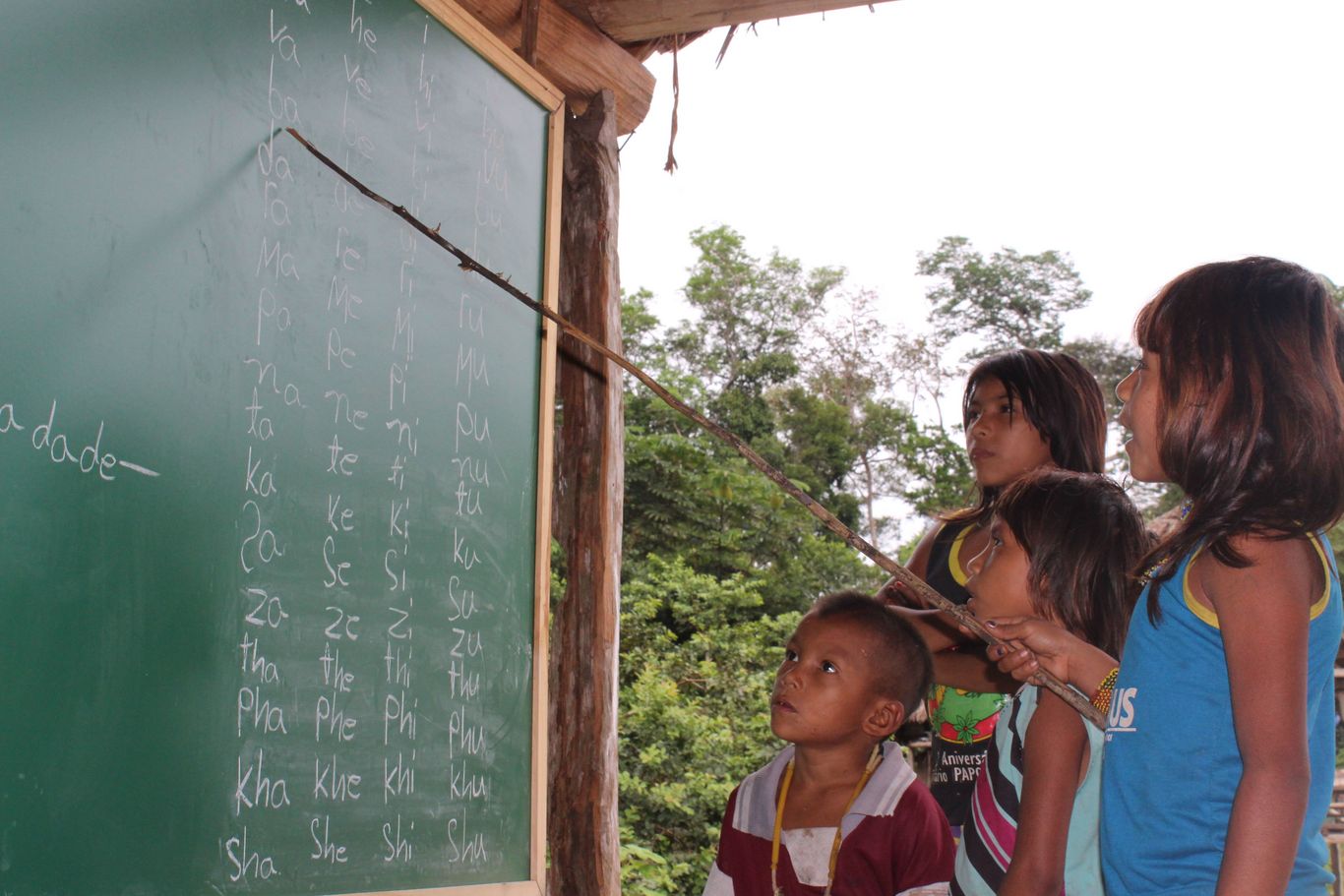 Kinder stehen vor einer Schultafel. Ein Mädchen zeigt mit einem Stock auf die geschriebenen Laute.