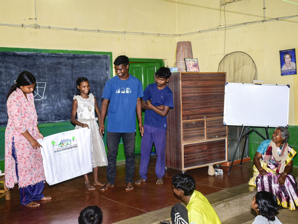 Mehrere Kinder präsentieren gemeinsam ein Plakat in einem Klassenraum, eine Betreuerin sitzt daneben und hört zu.