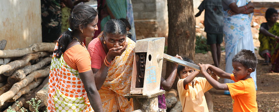 Zwei Frauen in Sari stehen bei einem Wasserbrunnen und 2 Kinder pumpen mit dem Hebel.