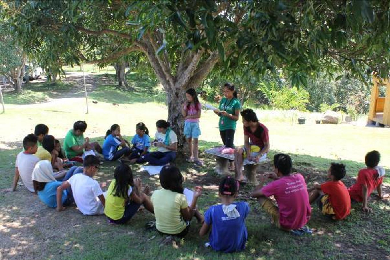 Kinder sitzen im Kreis unter einem Baum und lauschen einer Betreuerin von CAM