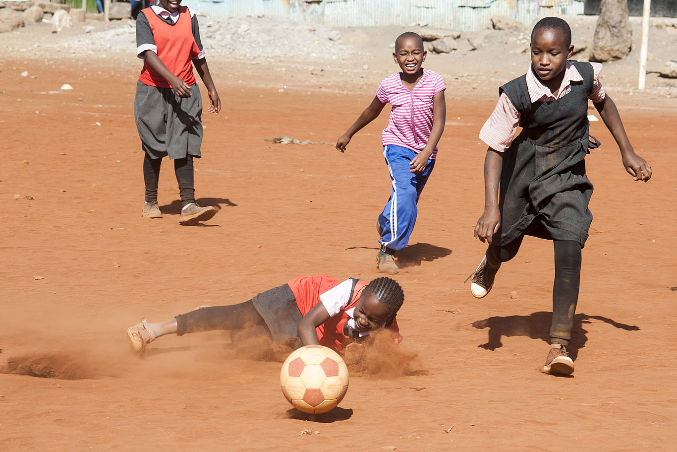 Mehrere Mädchen spielen auf rotem Boden Fußball, eine ist gestürzt und lacht.
