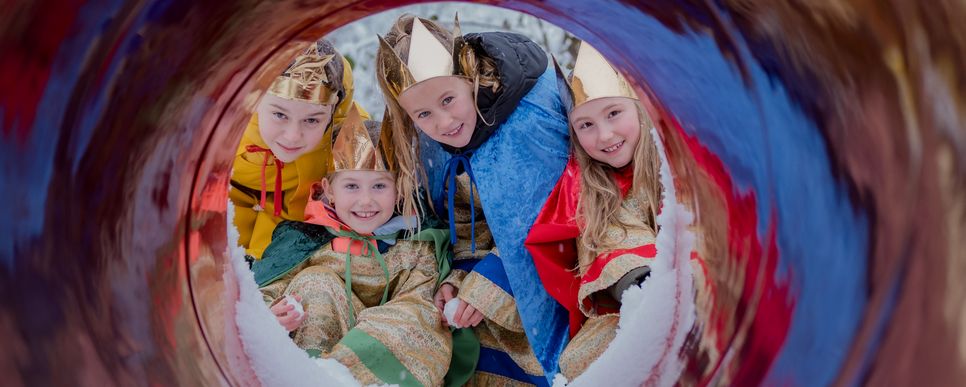 Vier Kinder, in Kleidung der heiligen drei Könige samt Sternsinger, schauen durch ein Rohr in die Kamera.