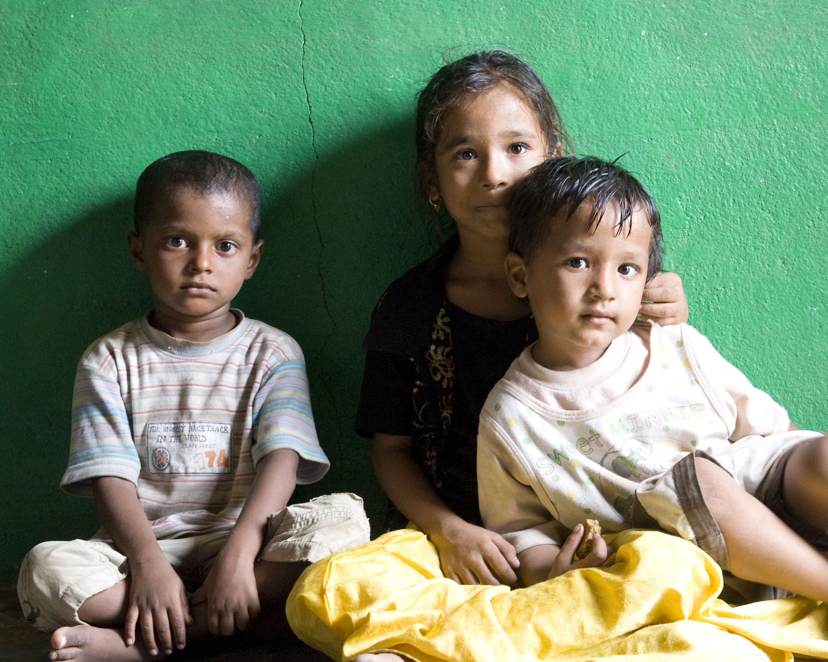 Drei Kinder aus Indien sitzen vor einer grünen Wand am Boden.