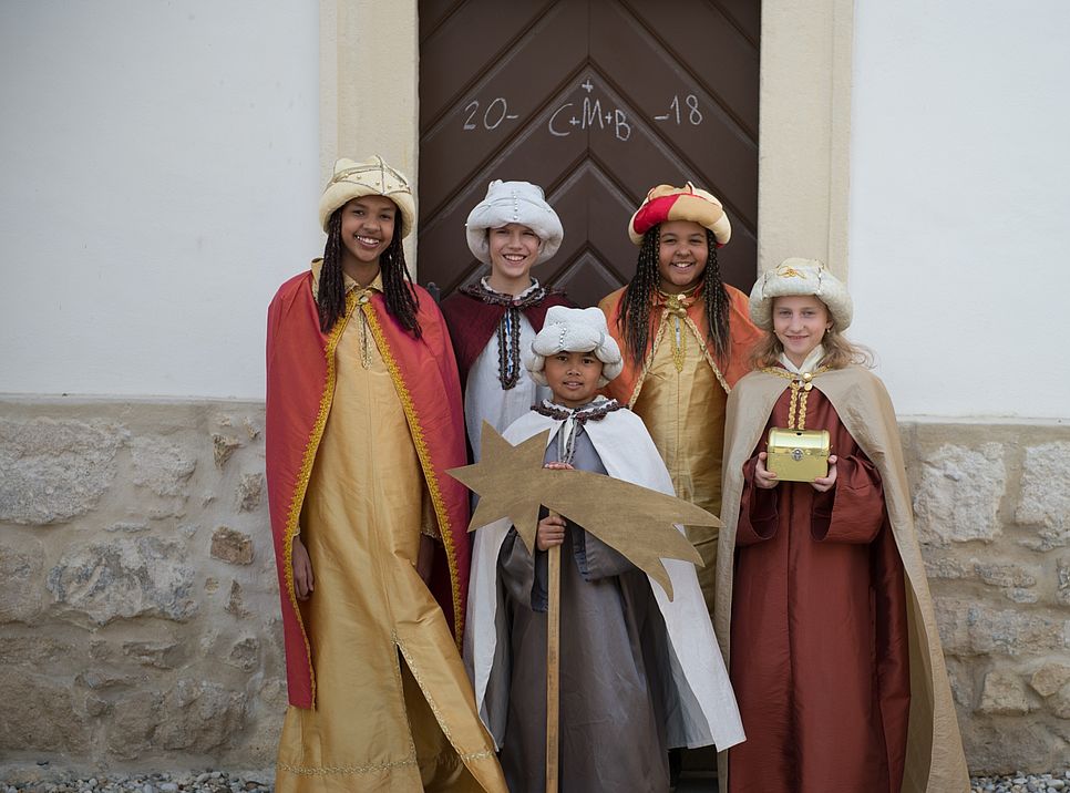 Fünf Sternsinger/innen stehen vor einer Tür mit Haussegen.