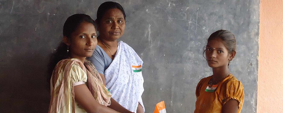 Zwei Frauen in Saris und ein Mädchen stehen in einem Klassenzimmer und blicken in die Kamera.