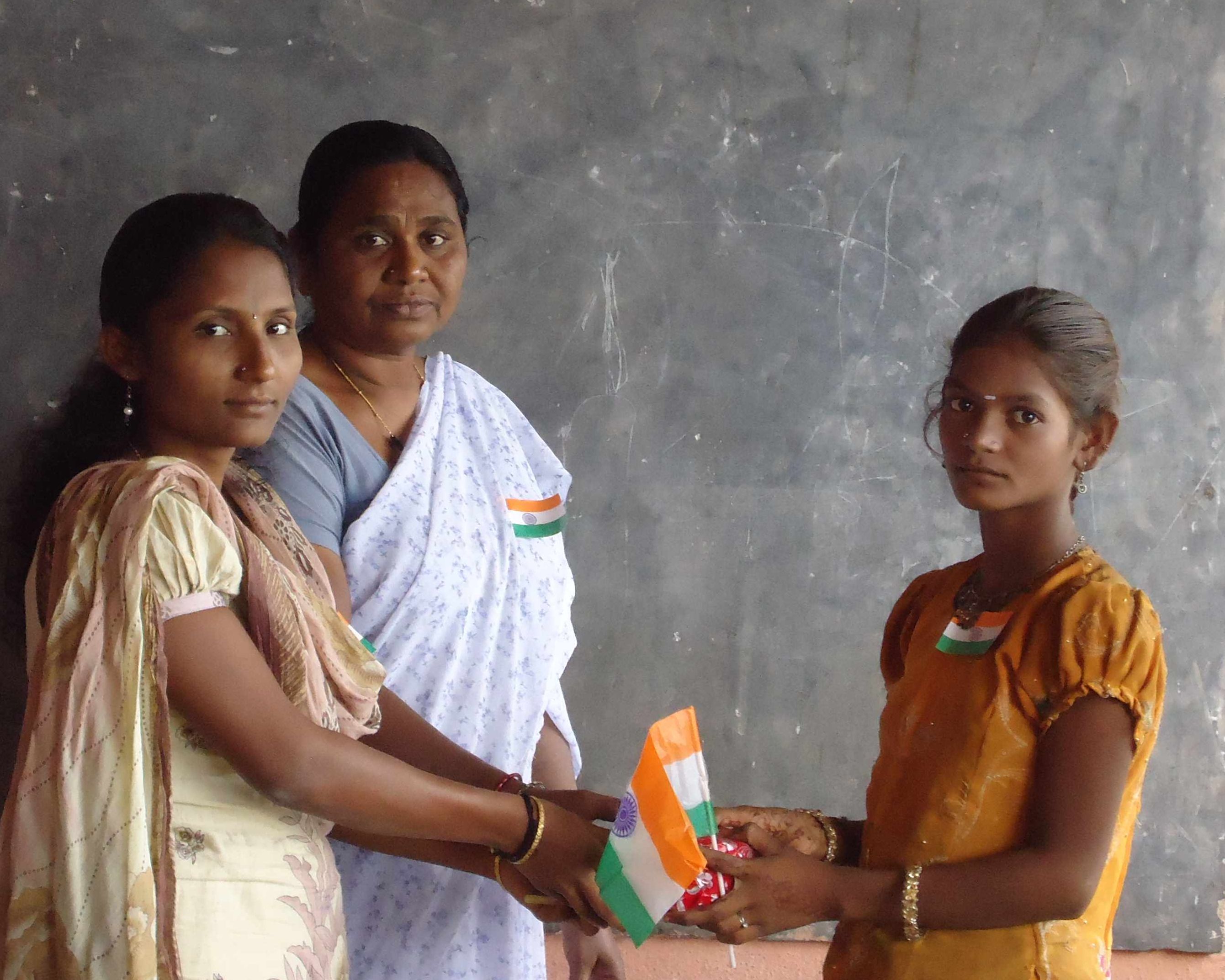 Zwei Mädchen stehen vor einer Schultafel und einer Frau. Die Mädchen überreichen sich ein Geschenk, eine davon hat eine indische Fahne in der Hand.