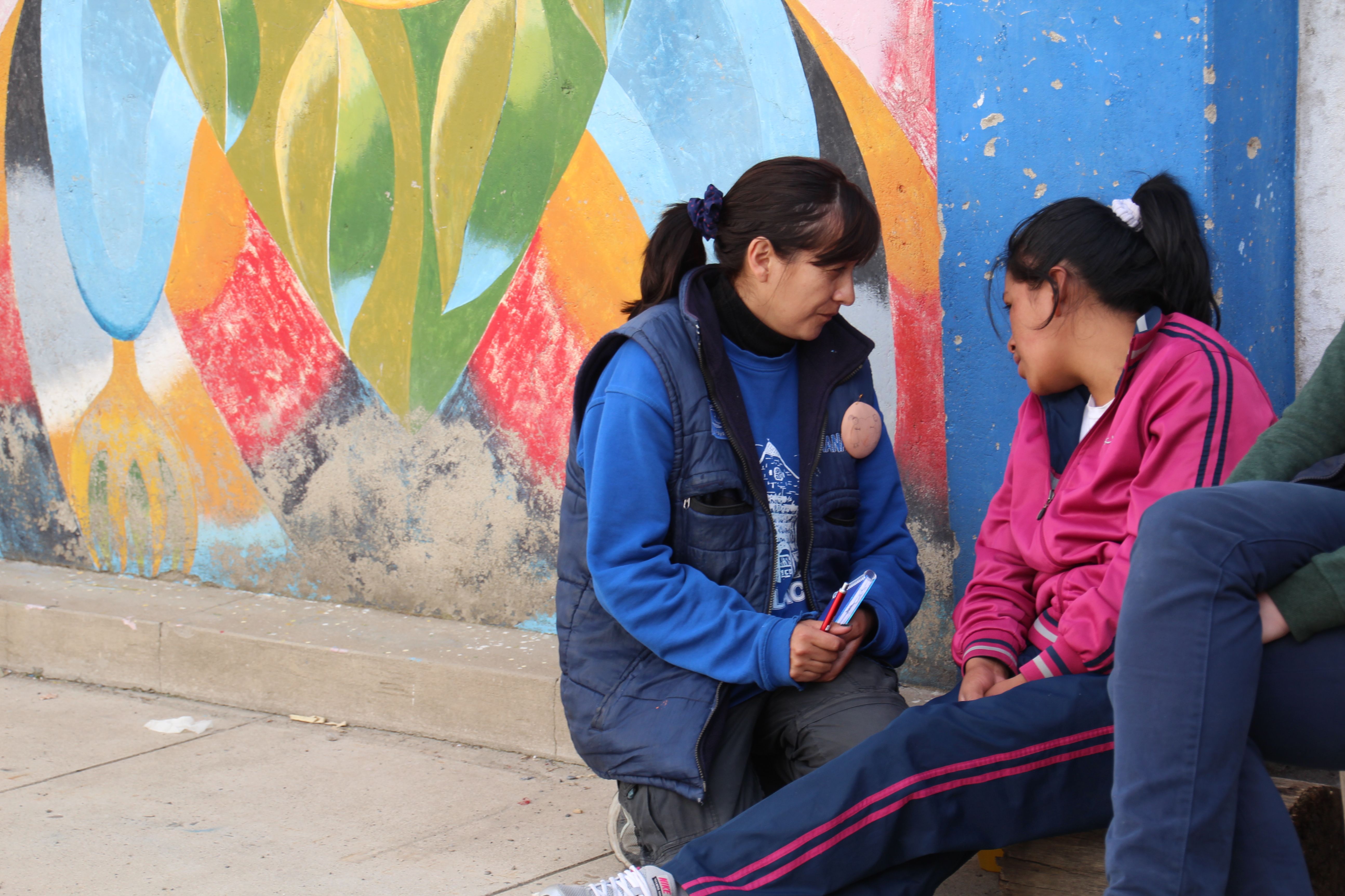 Eine Frau kniet neben einer jungen Frau die auf der Straße sitzt. Beide sind sich zugewandt und sprechen miteinander.