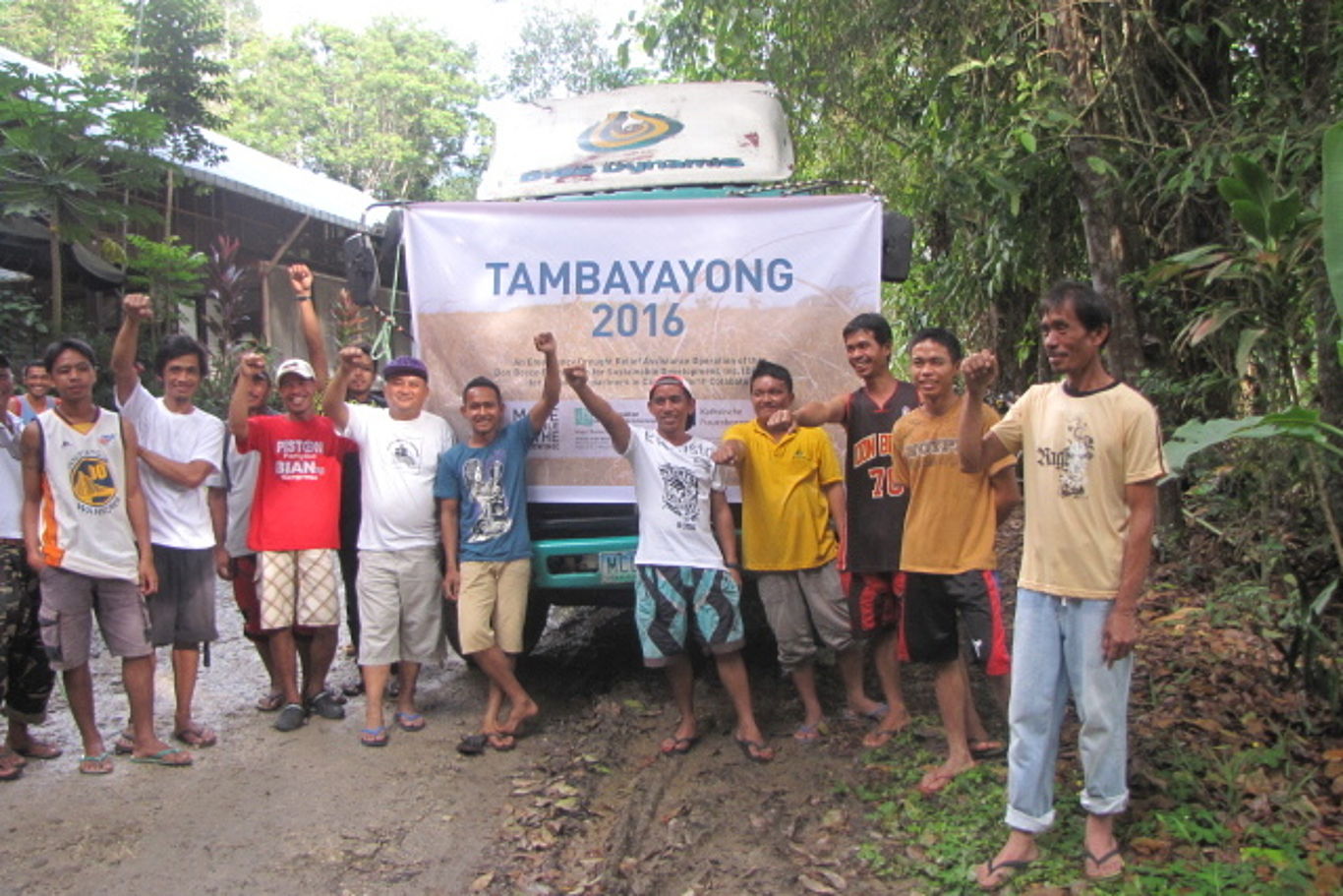 Eine Gruppe Männer und Jugendlicher steht vor einem Transparent mit der Aufschrift "Tambayayong" (übersetzt: Zusammenarbeit)