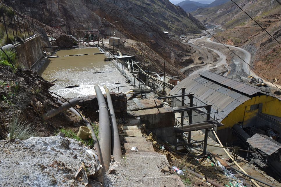 Das Bild zeigt eine industrielle Abwasseranlage in einem Bergbaugebiet, eingebettet in eine trockene Gebirgslandschaft. Zu sehen sind verschmutzte Wasserbecken, Rohre und verfallene Strukturen. Der Eingriff in die Natur und mögliche Umweltbelastungen sind deutlich erkennbar.