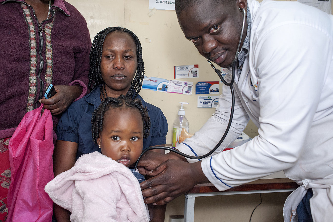 Die Gesundheitsklinik - im Bild Dr. Duncan Khaemba - bietet medizinische Behandlung für die Menschen. Ein Doktor hält sein Stethoskop an ein kleines Mädchen, welches auf seiner Mutter sitzt.
