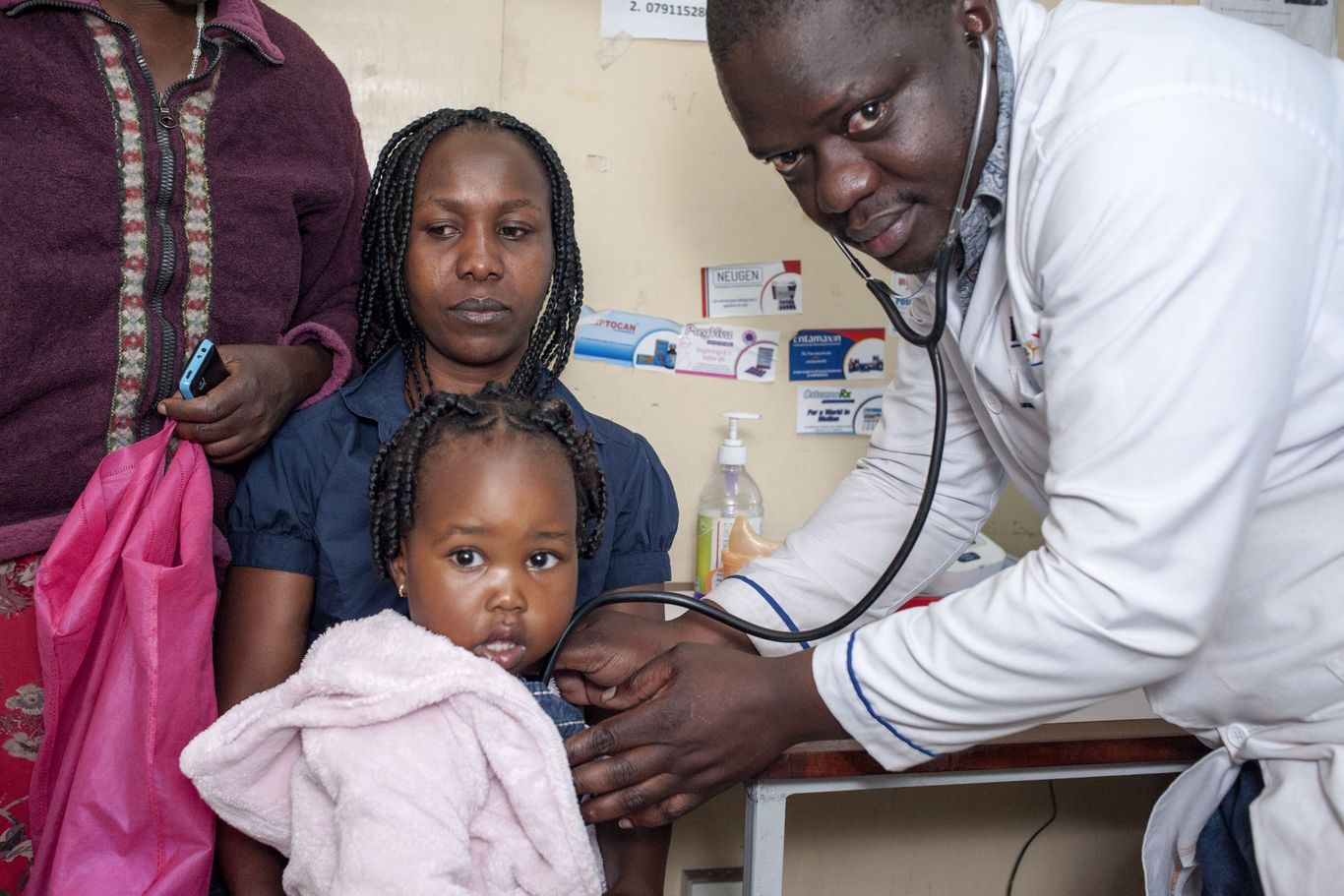 Ein Doktor hält sein Stethoskop an ein kleines Mädchen, welches auf seiner Mutter sitzt.