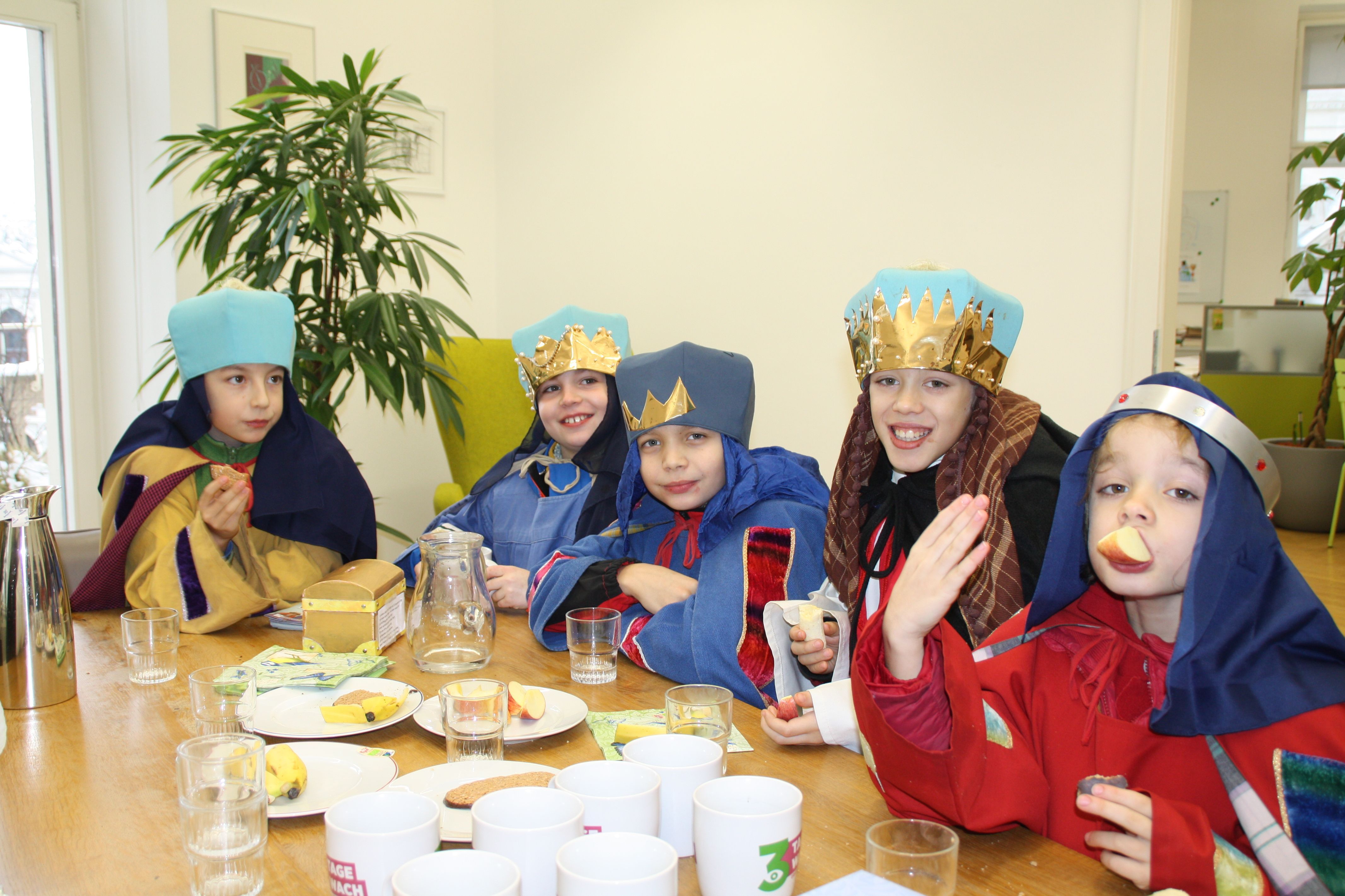 Eine Gruppe aus fünf Sternsinger Jungen sitzt an einem Tisch und stärken sich mit Obst und Keksen. Auf dem Tisch steht ihre Spenden-Kassa.