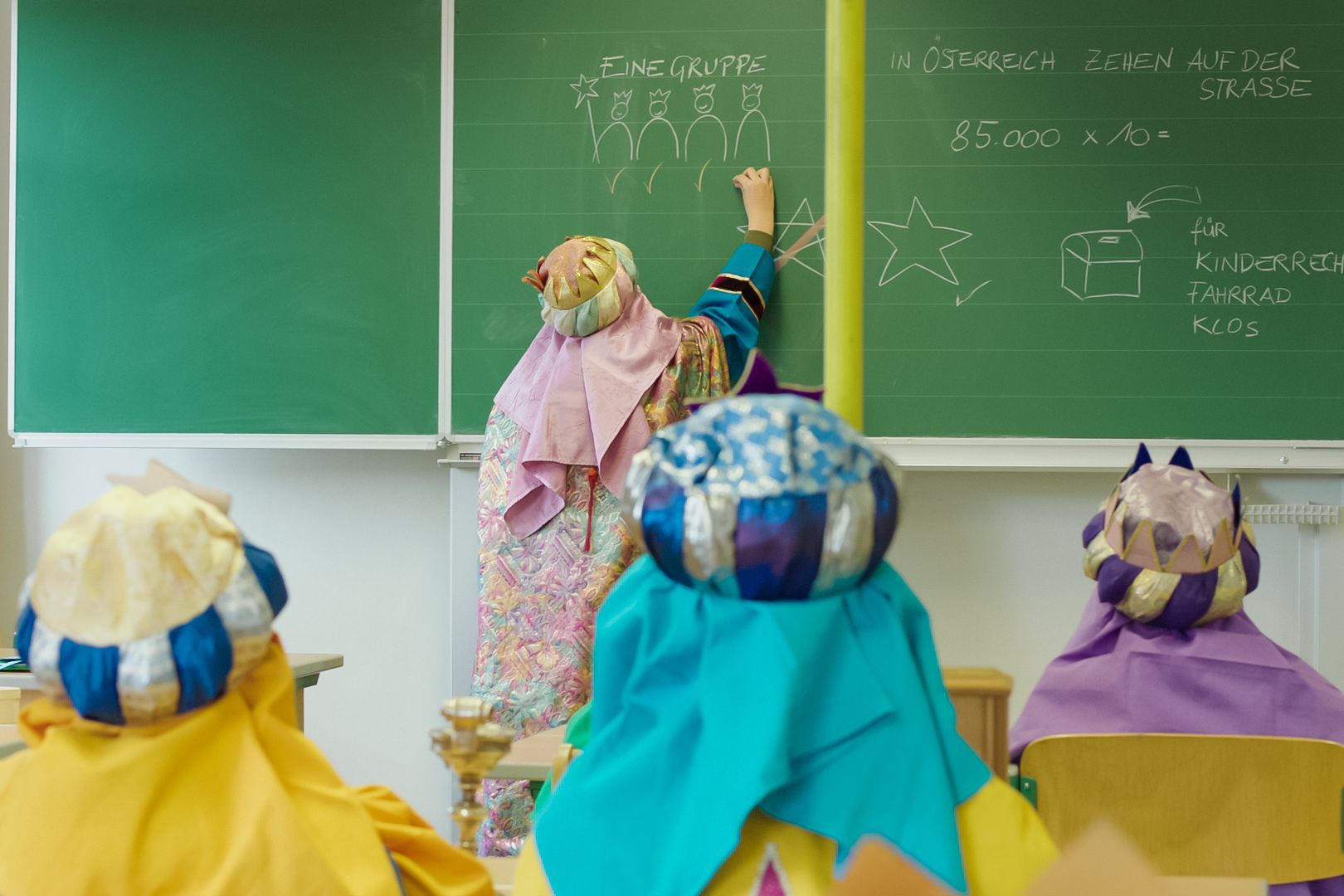 Ein Kind malt und schreibt etwas auf die Tafel, andere Kinder hören gespannt zu. Alle Kinder tragen Kleidung und Kronen der heiligen drei Könige.