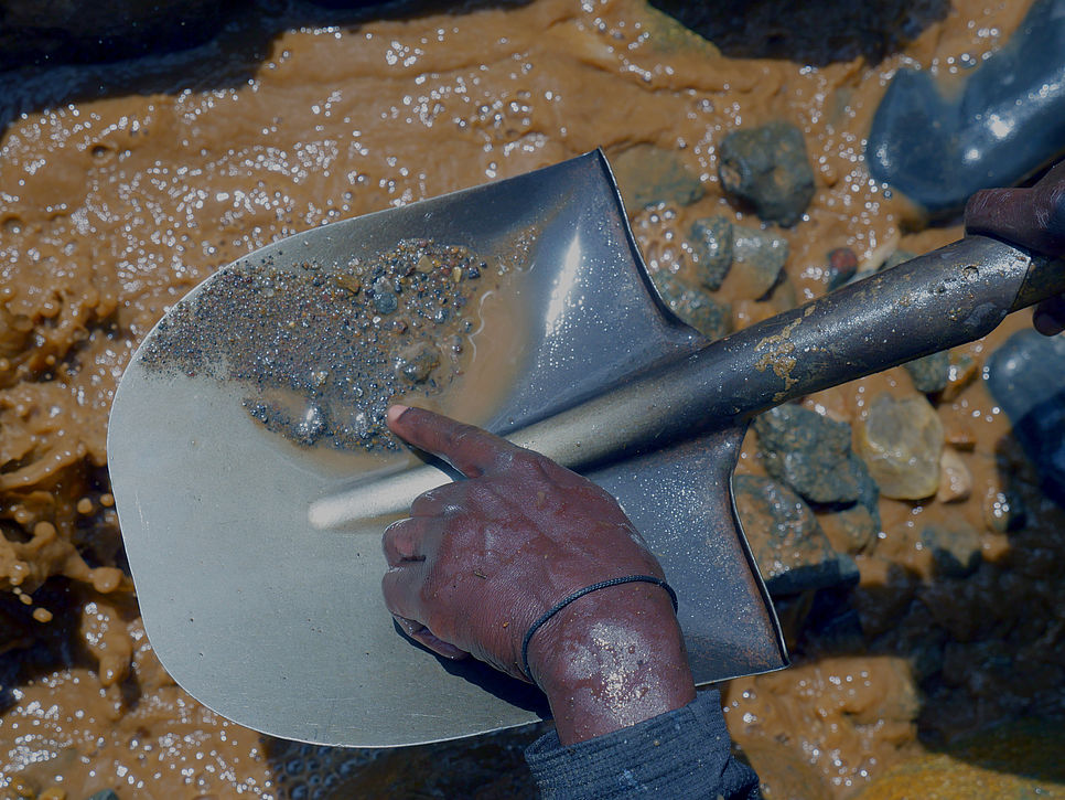 eine Hand zeigt auf eine Schaufel, auf der sich kleine Metallstücke befinden und im Hintergrund sind Schlamm und Steine