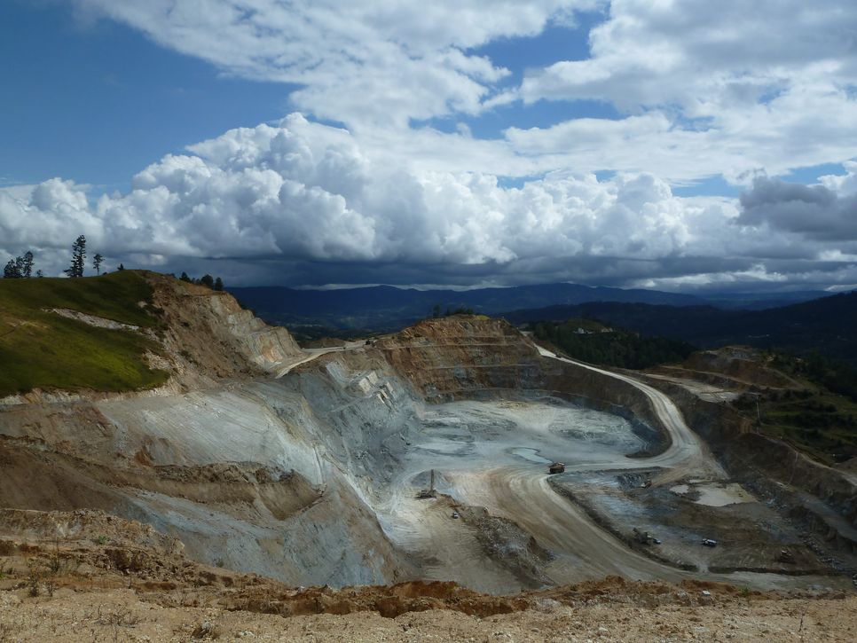 Das Bild zeigt eine große Goldmine im Tagebau in Guatemala, eingebettet in eine hügelige Landschaft. Der Boden ist tief ausgehoben, das Gestein wirkt karg und grau, einzelne Baufahrzeuge sind erkennbar. Über der Szene ziehen dichte Wolken am Himmel auf.