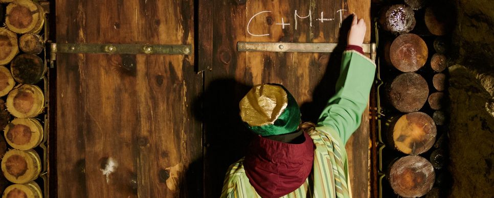 Foto: Weilguny Man sieht einen Sternsinger von hinten wie er das CMB auf eine Holztür schreibt.