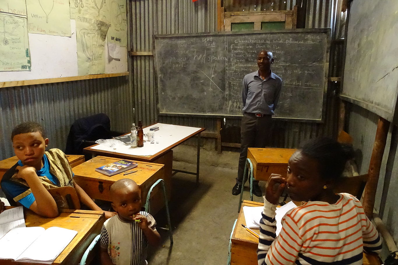 Kinder sitzen in einem Klassenraum von MPC, vor der Tafel steht ein Lehrer.