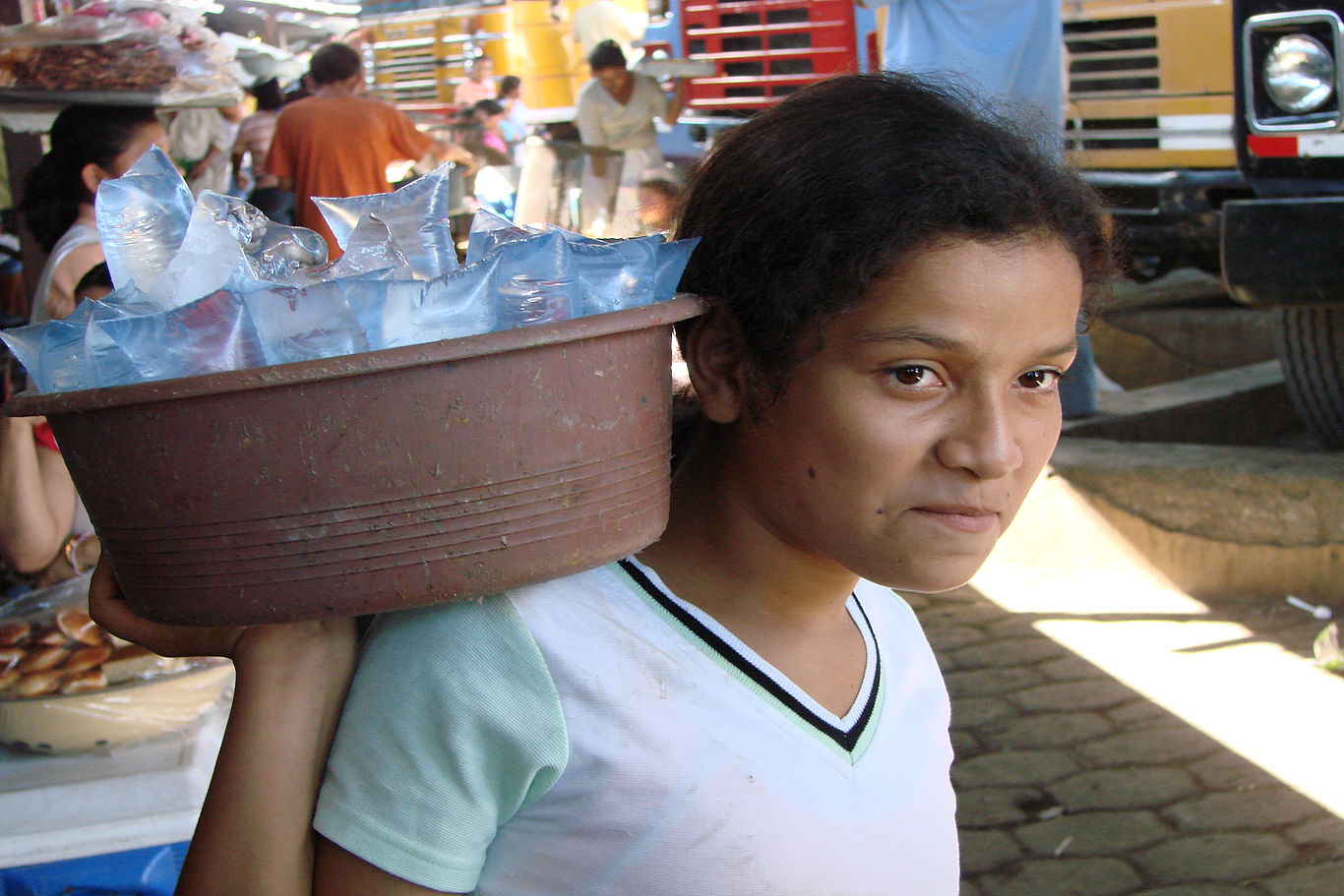 Junge Straßenverkäuferin trägt einen Korb mit Wasserflaschen auf der Schulter.