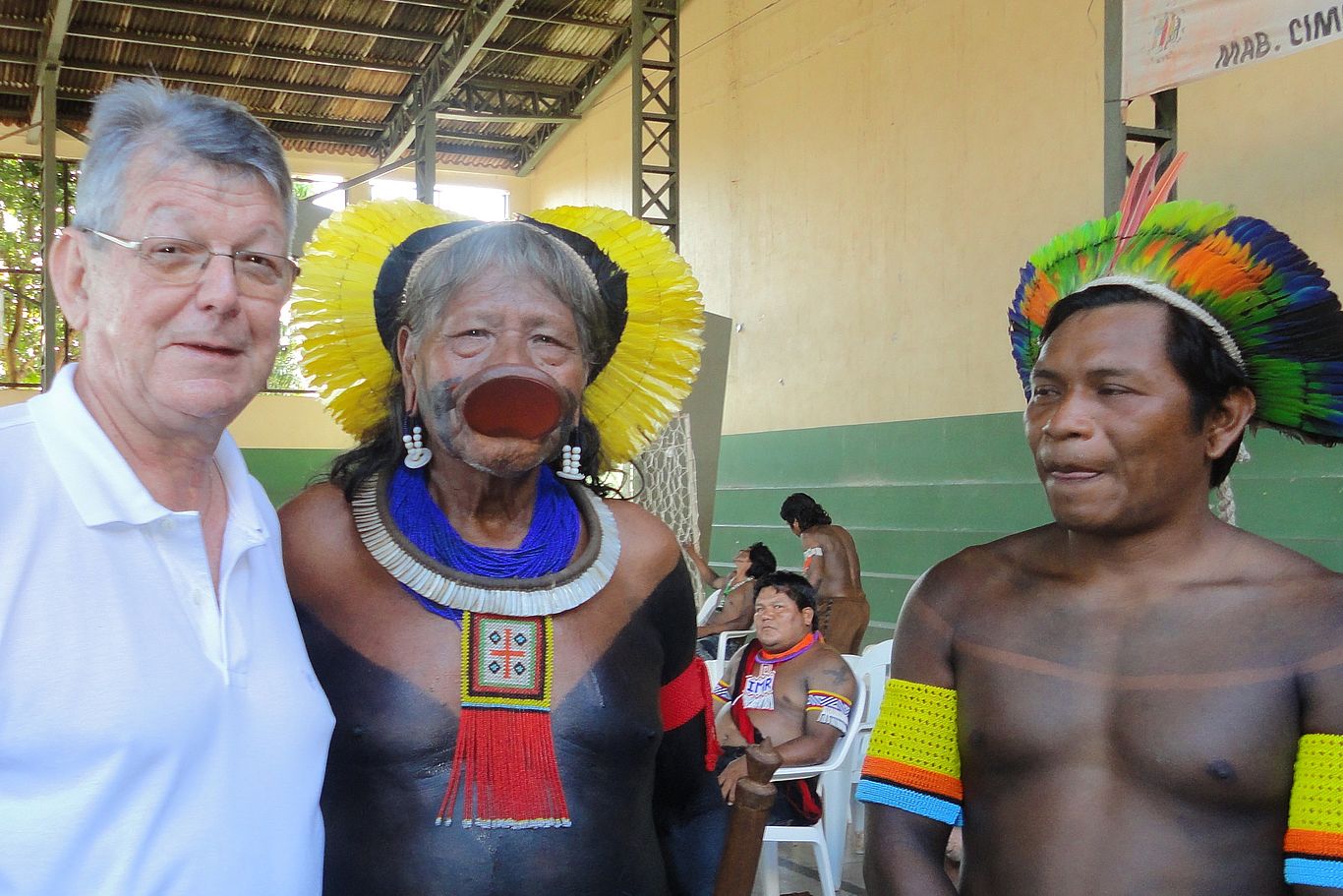 In Brasilien steht die katholische Kirche, z.B. Alt-Bischof Erwin Kräutler, den indigenen Völkern zur Seite. Drei Männer, zwei davon mit Federn geschmückt, schauen in die Kamera
