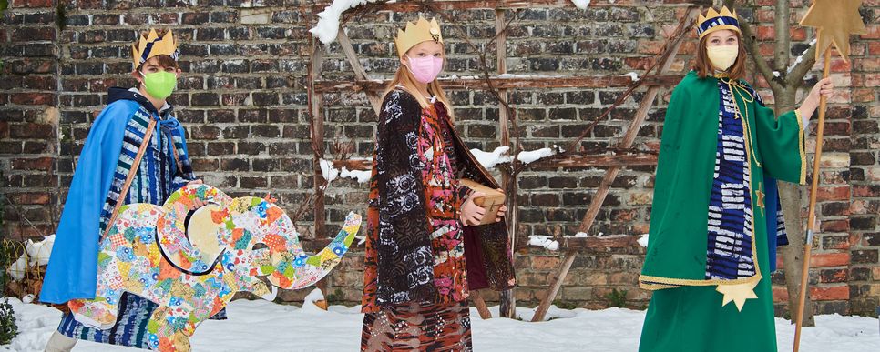 Foto: Paul Feuersänger Drei verkleidete Sternsinger_innen mit Masken gehen im Schnee vor einer Ziegelwand. Ein Sternsinger hat einen bunten Pappelefanten dabei. Eine Sternsingerin trägt den Stern.