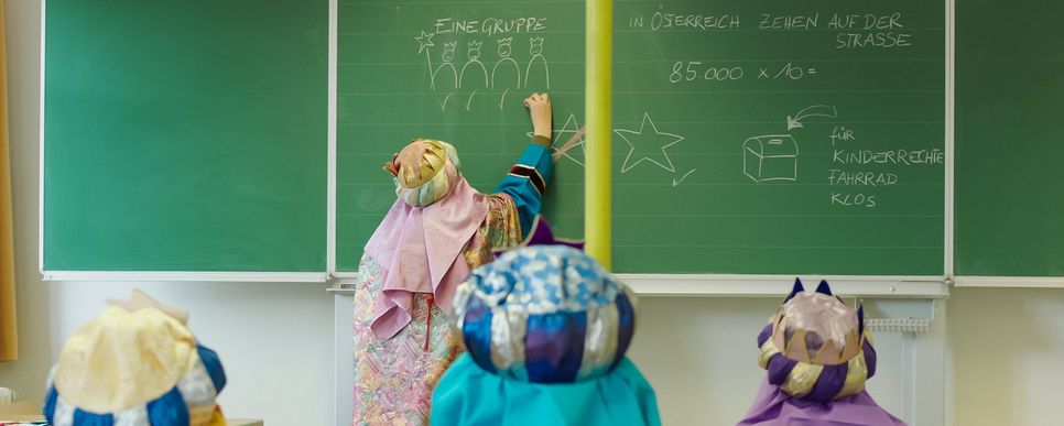 Ein Kind schreibt Informationen zur Sternsingeraktion auf eine grüne Tafel und drei andere Kinder sitzen auf Stühlen und schauen zu.