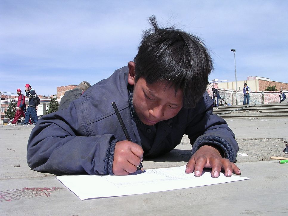 Ein Bub schreibt auf einem Zettel, welcher auf der Straße liegt