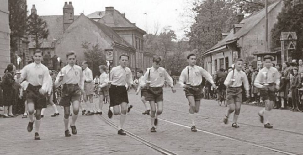 Lichtstafette: Sieben Buben laufen mit Fackeln in der Hand auf Straße, links und rechts von ihnen Spalier.