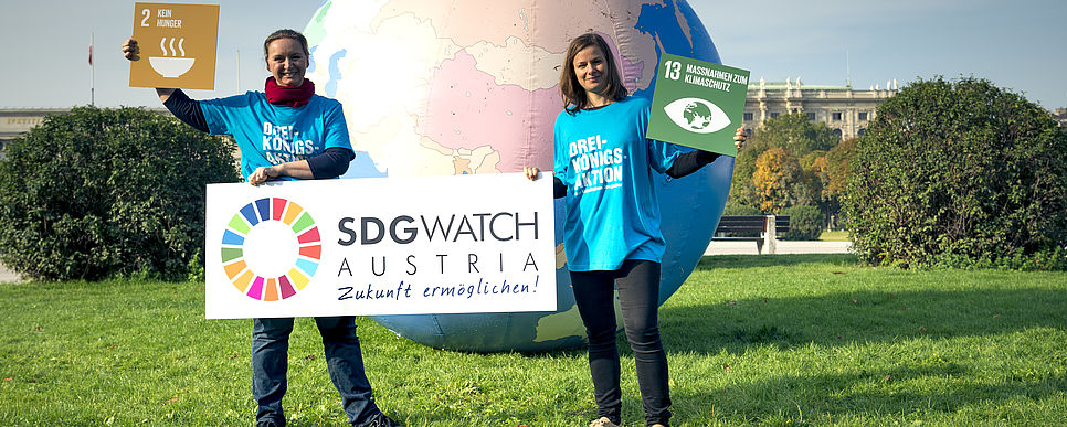 Aufruf zur Umsetzung der SDGs Zwei Frauen stehen hinter einem großen Weltkugel Ball auf einer Wiese und halten ein Transparent und SDG Ziele in die Luft