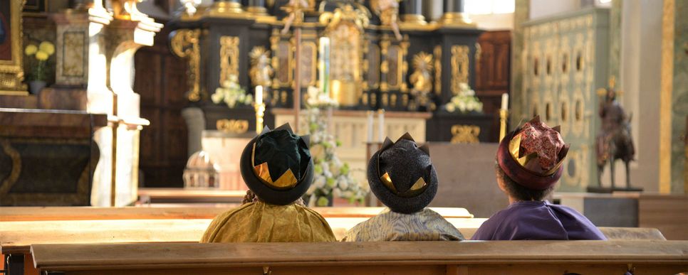 Gottesdienst mit den Heiligen Drei Königen feiern. Drei Könige sitzen in einer Kirchenbank und schauen auf den Altar.