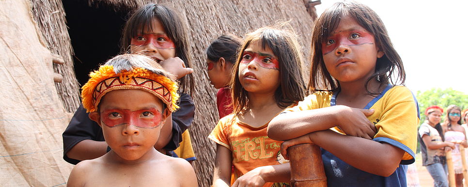 Indigene Völker in Brasilien, Foto: Herret Kinder mit Gesichtsbemalung stehen vor einer Strohhütte