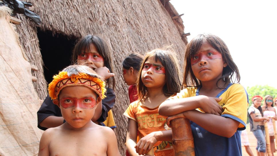 Vier indigene Kinder aus Brasilien stehen draußen vor einer Hütte. Die Augen sind mit einem Band aus roter Farbe umzogen.