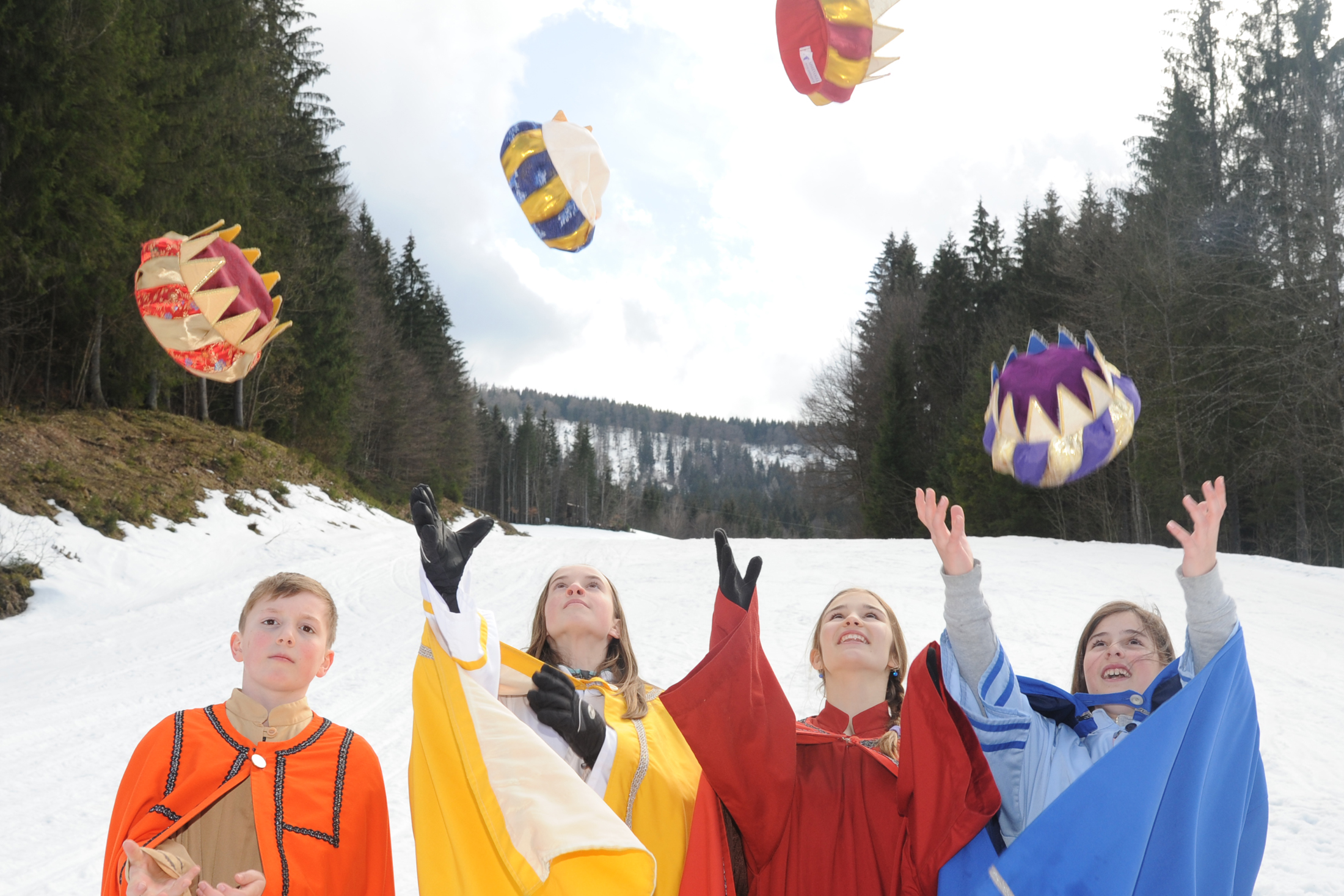 Eine Gruppe von Sternsingern, drei Mädchen und ein Junge, stehen vor einem verschneiten Feld. Sie werfen ihre Kronen in die Höhe.